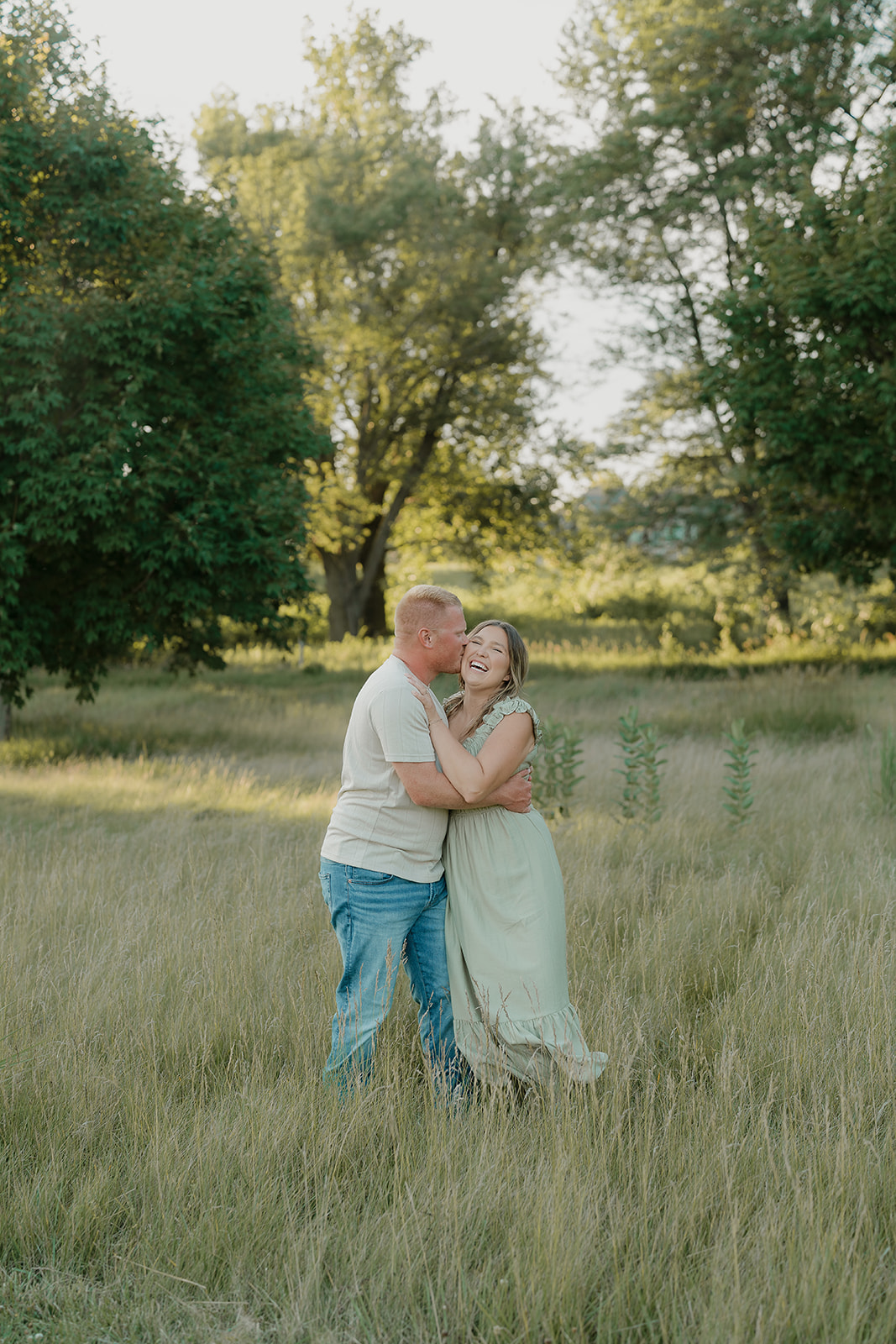 The groom kisses his fiancée on the cheek as she throws her head back laughing, both standing in tall golden grass with trees glowing behind them in summer engagement photo outfits
