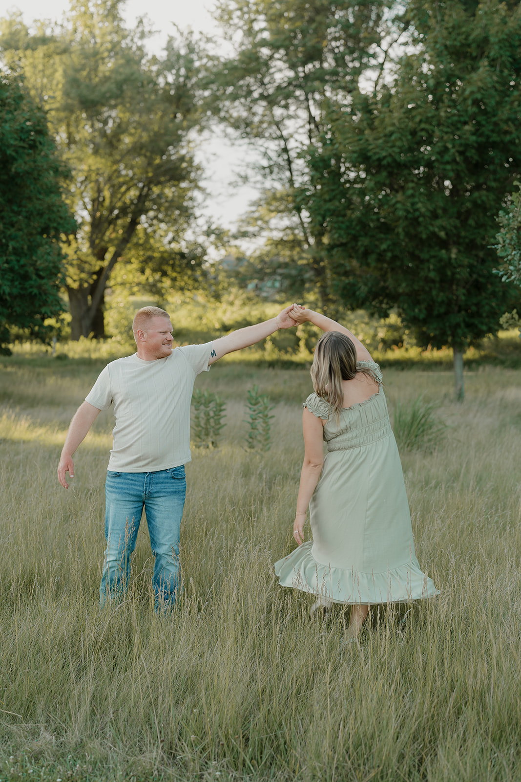 The man spins his fiancée in a summer engagement photo outfit — a sage off-shoulder dress — through tall golden grass at Ada Hayden Heritage Park in Iowa
