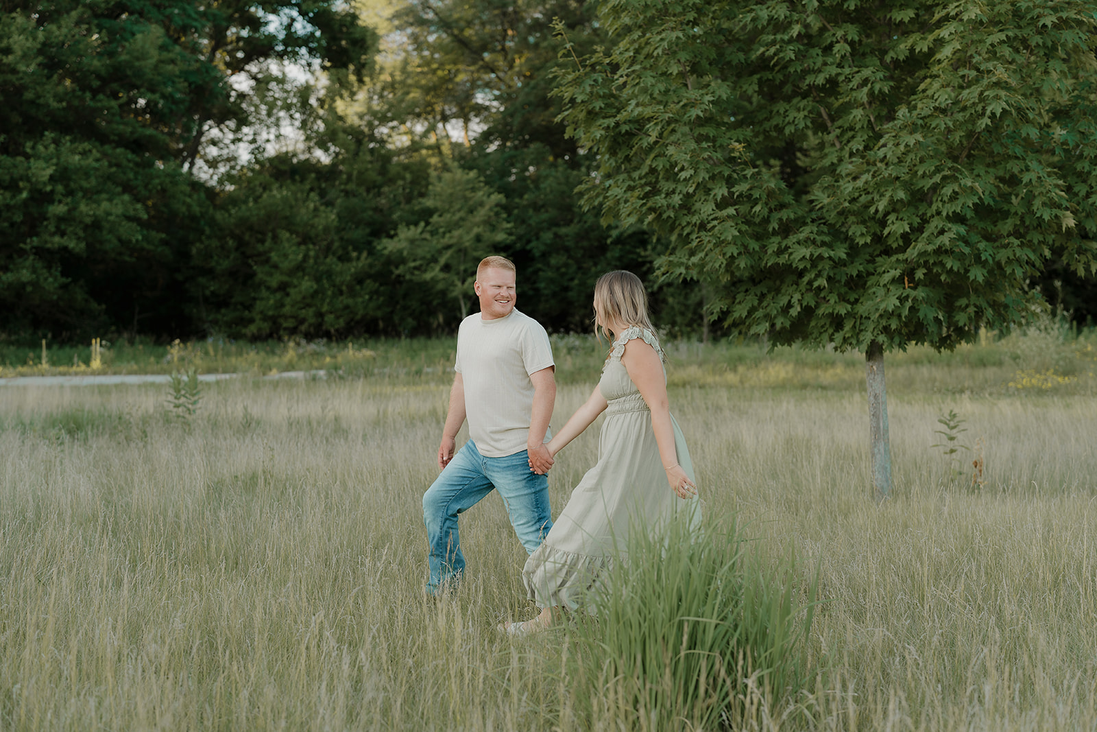A couple holds hands and walks through a tall grass meadow at Ada Hayden, the woman leading the way in her sage ruffle dress as her partner smiles at her
