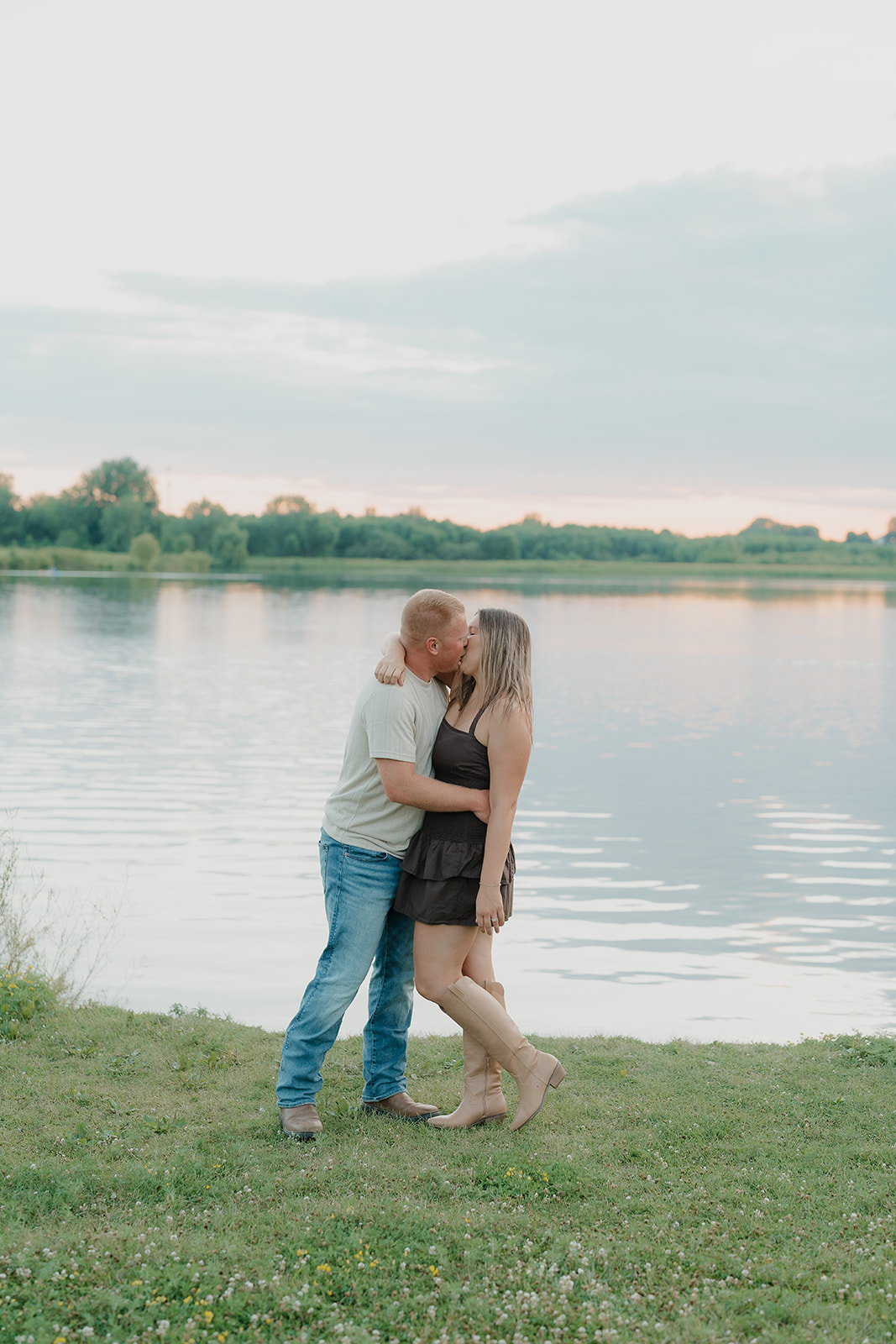 A couple in summer engagement photo outfits shares a kiss at the edge of Ada Hayden lake at sunset, her dark smocked mini dress and tan knee-high boots paired with his jeans and tee
