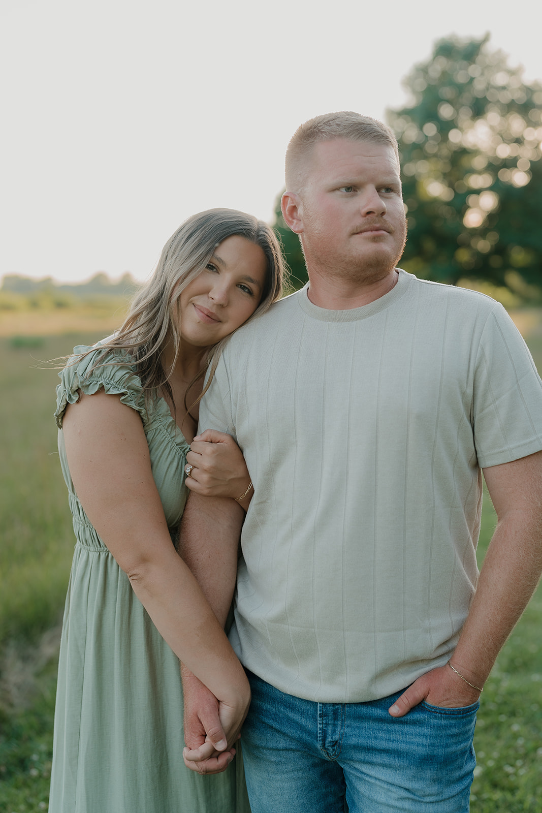 A close-up portrait of the woman resting her head on her partner's shoulder in a sage green dress, her engagement ring visible as they stand in a field at dusk
