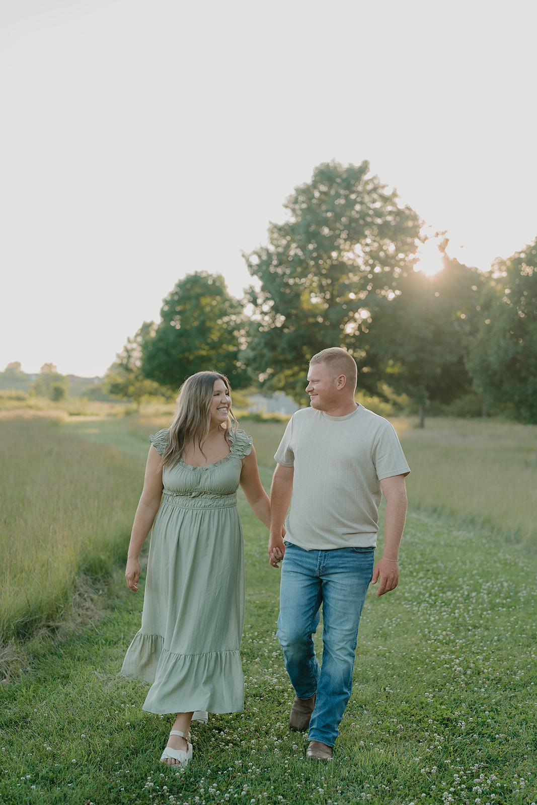 The couple walks hand in hand through a clover-filled meadow path at Ada Hayden, laughing together in the warm golden hour light
