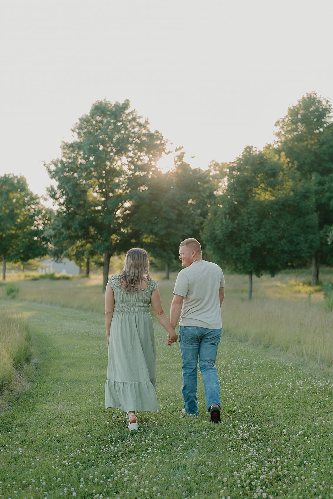 A couple wearing summer engagement photo outfits walks hand in hand away from the camera through a clover field toward golden sunlight filtering through the trees
