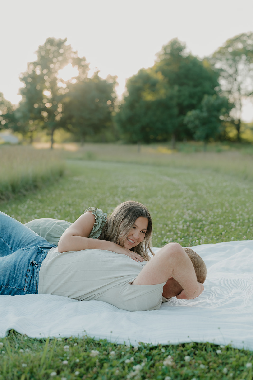 The couple lies together on a white blanket in a sunlit field, the woman resting on her partner's chest and smiling up at him with golden hour trees in the background
