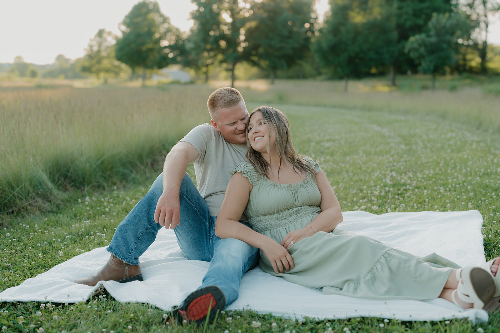 A couple in summer engagement photo outfits sits together on a white blanket in a clover field, the woman leaning back against her partner as he nuzzles her cheek
