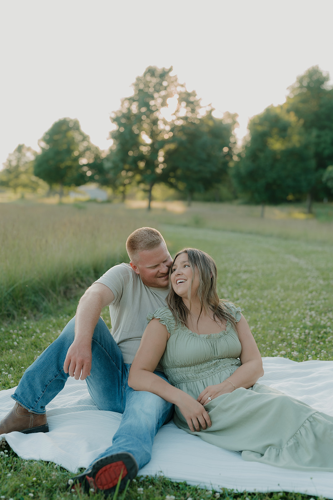 A couple relaxes together on a blanket in a clover field, the woman in a sage ruffle-strap dress leaning back and laughing as her partner nuzzles her cheek
