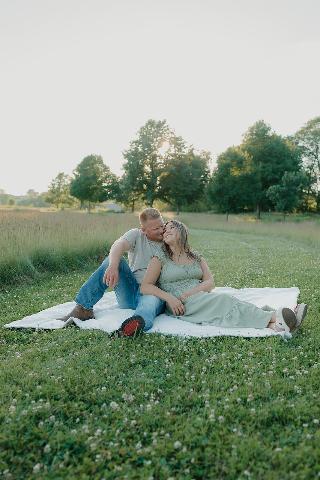 A couple sits together on a white blanket in a clover meadow at Ada Hayden, the man kissing her cheek as she smiles up at him in the warm summer light
