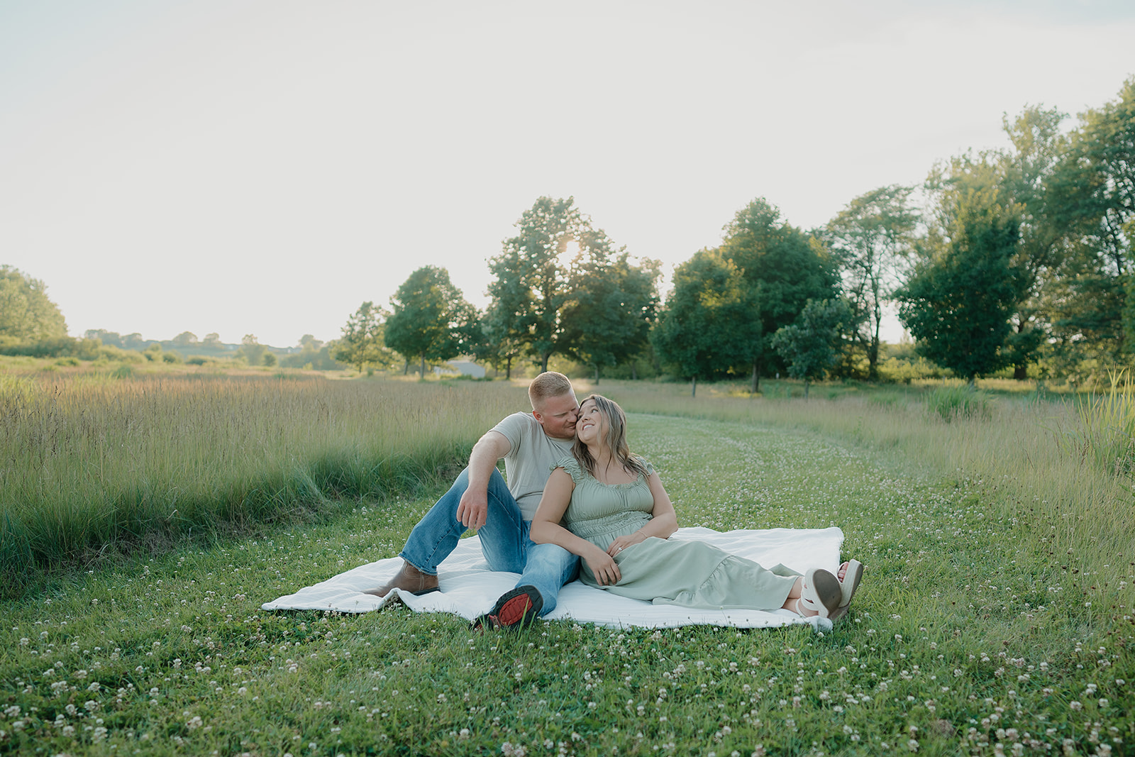 A couple sits together on a blanket in a clover meadow at Ada Hayden as the man kisses her cheek and she laughs up at him in the warm evening light
