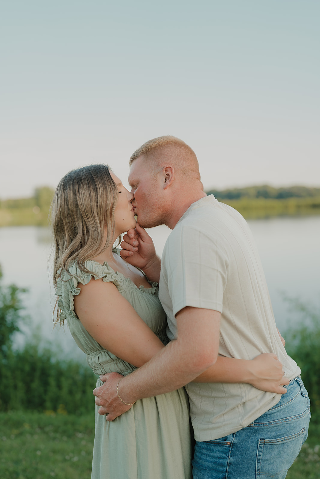 A couple shares a kiss near the water's edge at Ada Hayden in their summer engagement photo outfits — a sage ruffle dress and neutral ribbed tee with jeans

