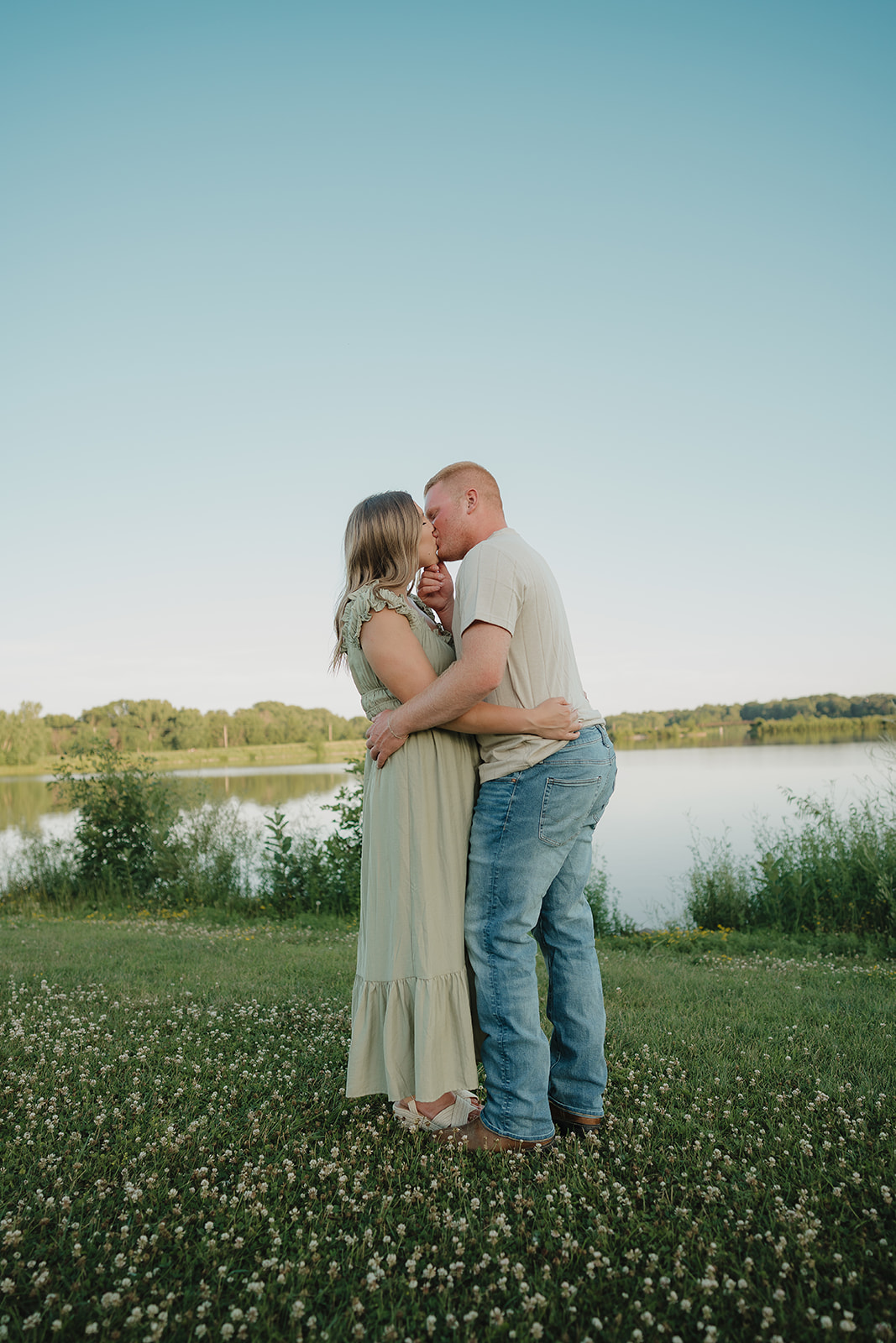 An engaged couple shares a kiss in a clover-filled field at Ada Hayden, Iowa, with a still lake and clear blue sky stretching behind them

