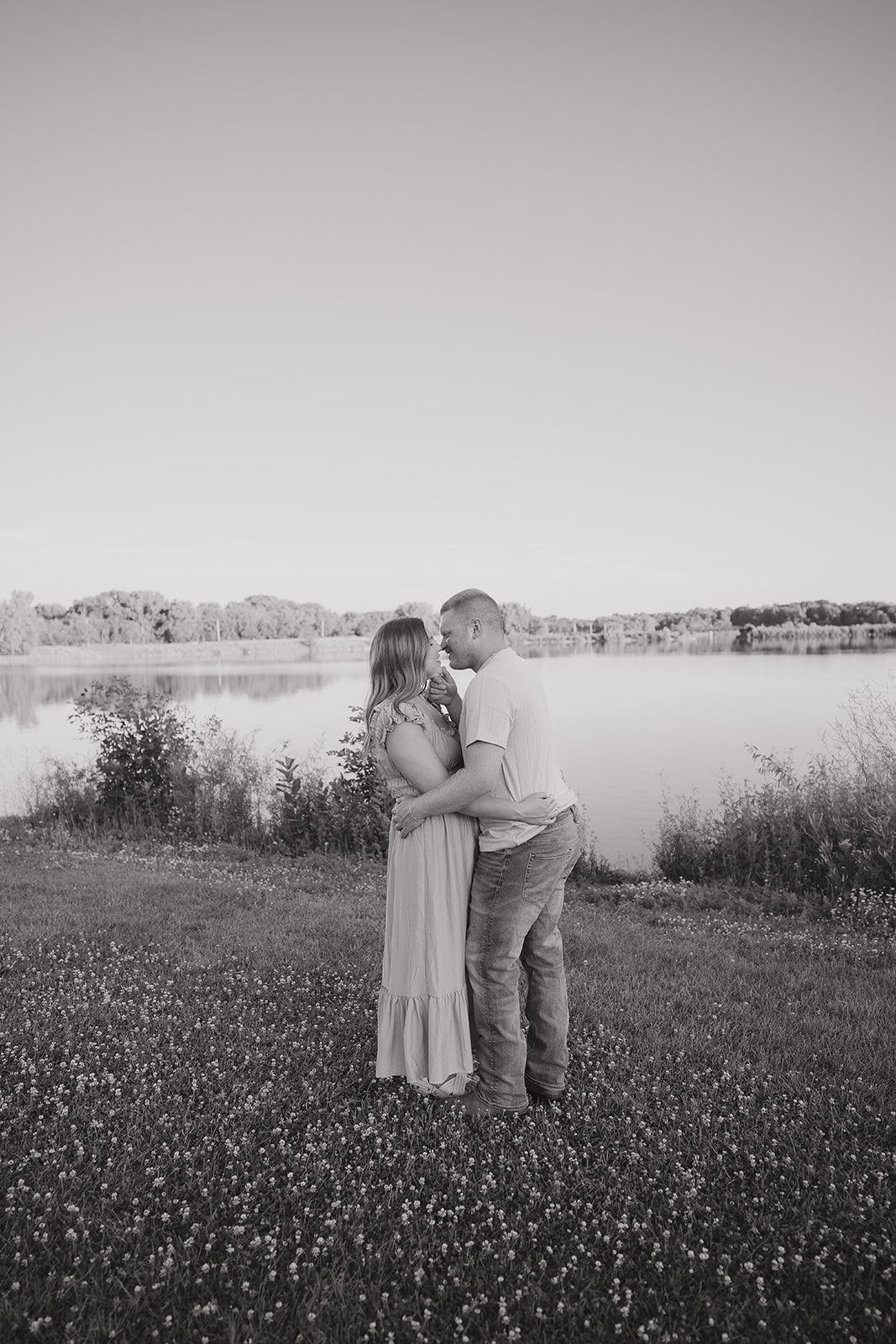 A black and white photo of the couple gazing at each other in a clover field at Ada Hayden, the still lake and treeline reflected softly behind them
