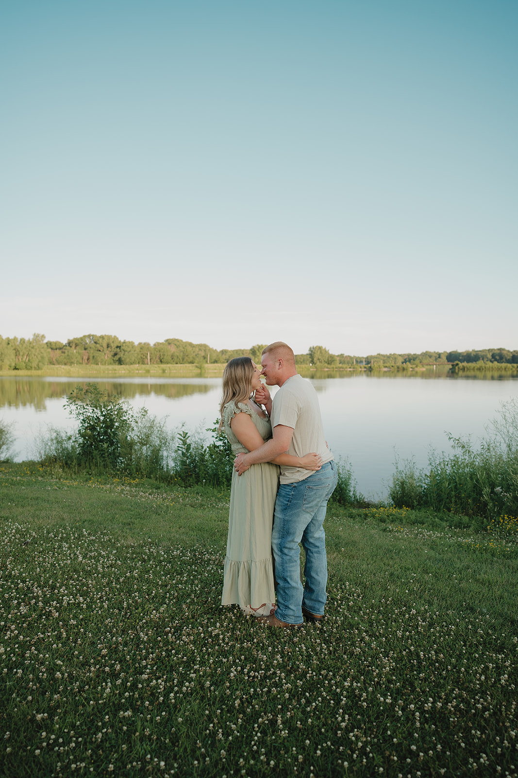 An engaged couple in summer engagement photo outfits — a sage green maxi dress and neutral tee with jeans — share a close moment beside a calm lake at Ada Hayden in Iowa

