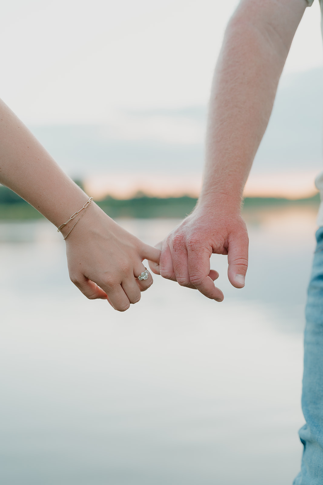 A close-up detail shot of the couple loosely holding fingertips, the woman's oval diamond engagement ring and gold chain bracelet visible against the soft lake backdrop
