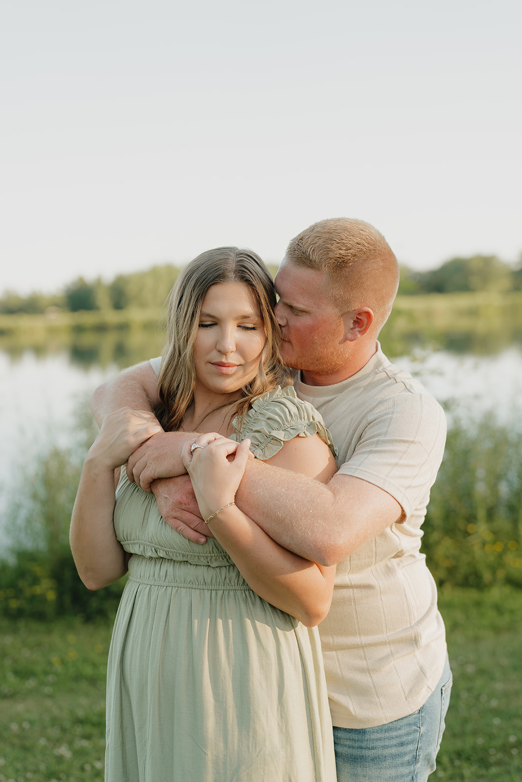 The groom wraps his arms around his fiancée from behind, kissing her cheek as she laughs and shows off her engagement ring beside the lake at Ada Hayden
