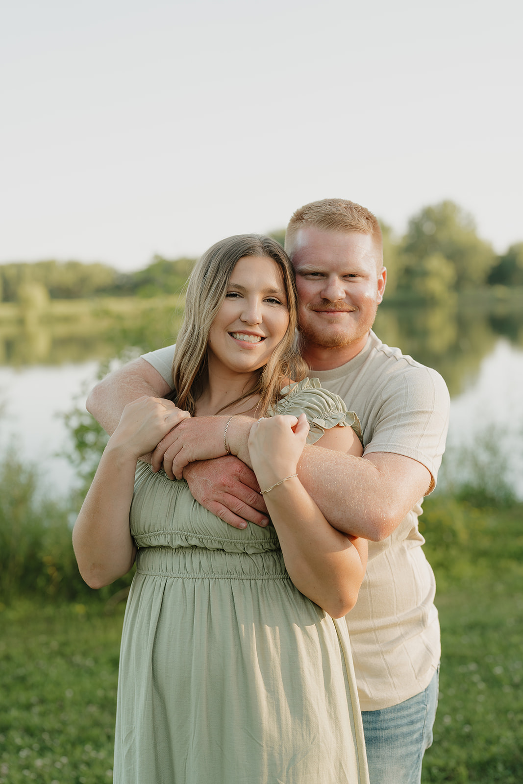 An engaged couple faces each other and smiles in front of lush green trees, the woman's arms resting on her partner's shoulders during their Iowa engagement session
