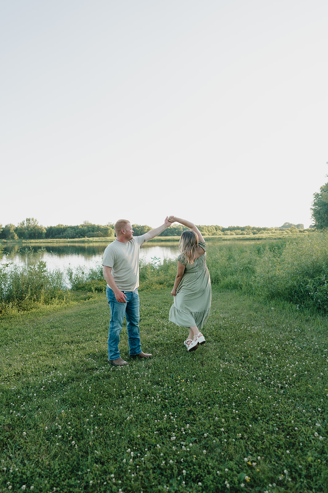 A couple in summer engagement photo outfits twirl together on the grass beside a peaceful lake, her sage maxi dress and his jeans and tee perfectly coordinated

