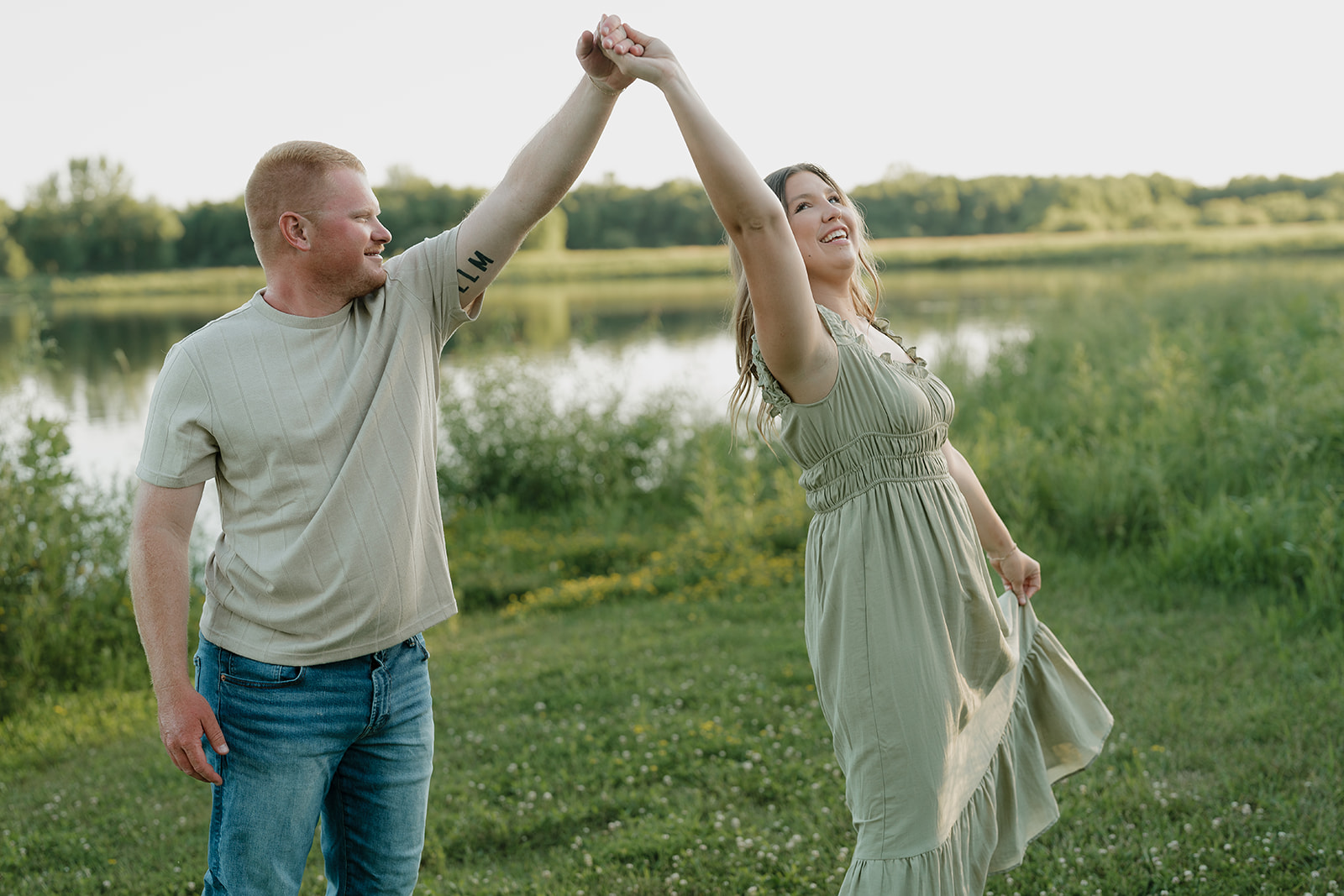 A couple wearing summer engagement photo outfits dance and spin together near the water's edge at Ada Hayden, the woman's sage green dress swirling in the golden light
