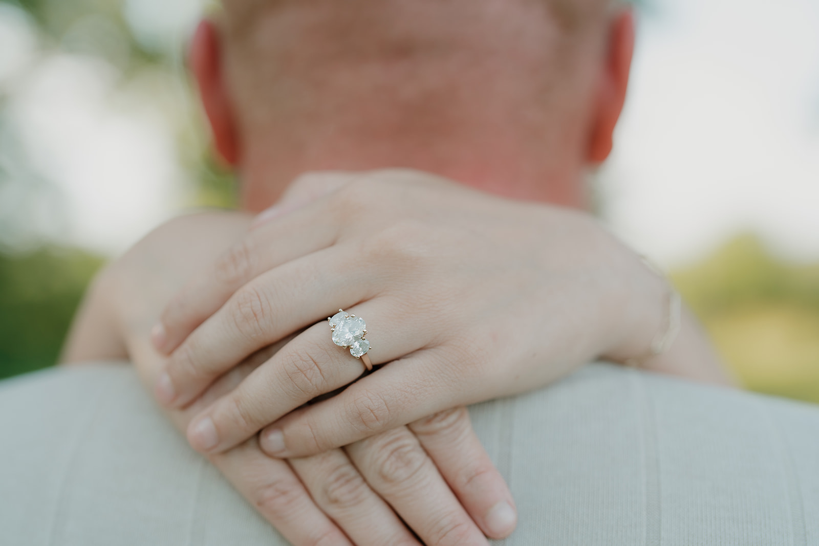 A close-up detail of the woman's three-stone oval diamond engagement ring resting on her fiancé's shoulder during their Ada Hayden engagement session

