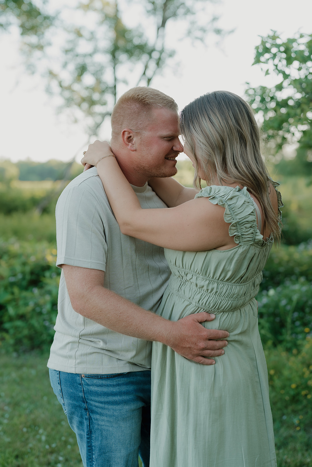 The couple stands forehead to forehead, both smiling, in summer engagement photo outfits among lush green wildflowers during their Ada Hayden session
