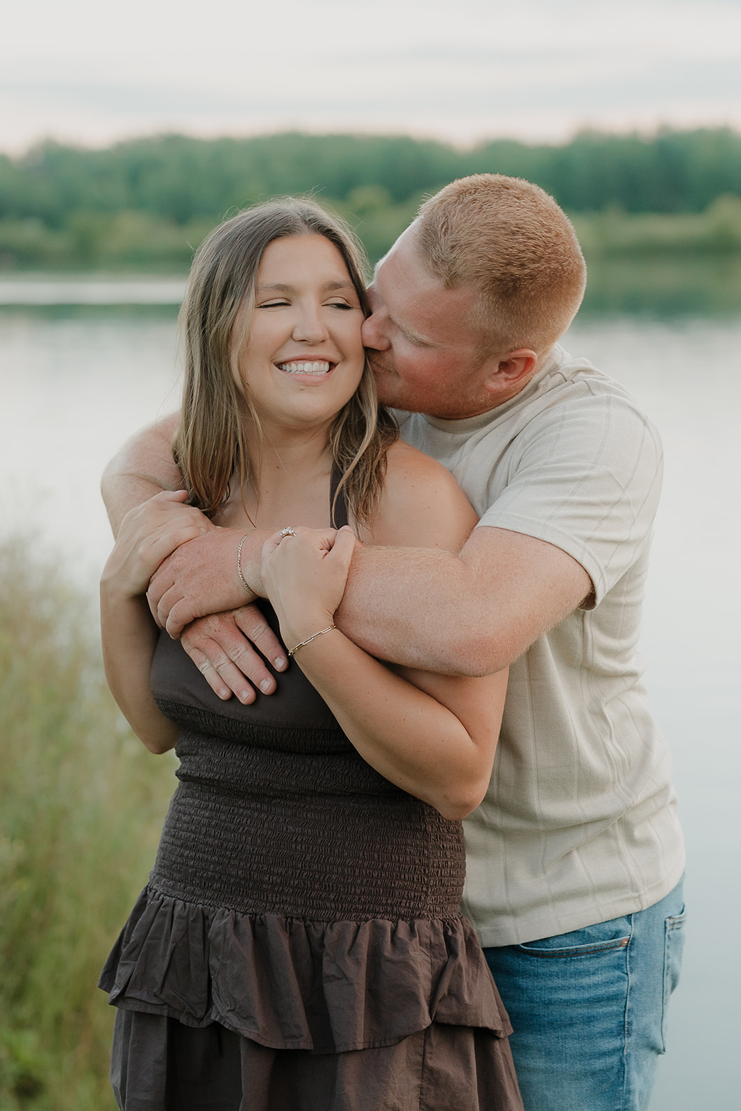 The groom wraps his arms around his fiancée from behind, kissing her cheek as she laughs and shows off her engagement ring beside the lake at Ada Hayden
