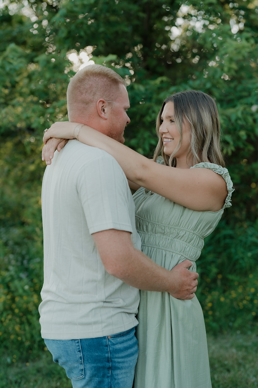 An engaged couple faces each other and smiles in front of lush green trees, the woman's arms resting on her partner's shoulders during their Iowa engagement session
