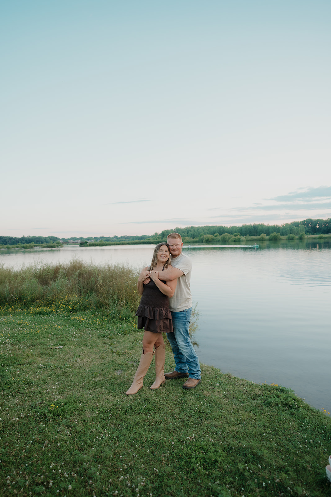 A couple embraces beside the calm water at Ada Hayden at dusk, the woman laughing in her dark mini dress and tan cowboy boots as her fiancé holds her from behind
