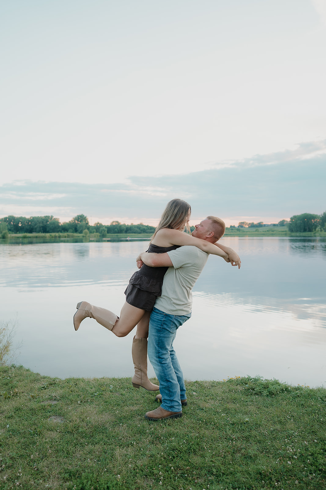 The man lifts his fiancée off the ground in a joyful embrace at the edge of Ada Hayden lake, both laughing against a pastel pink and blue sunset sky
