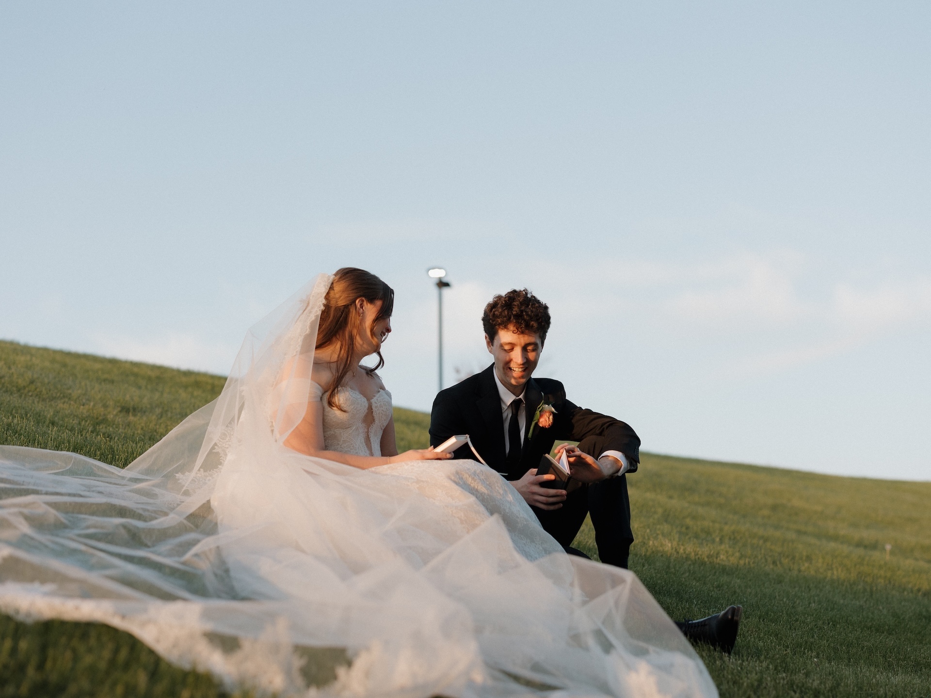 The bride and groom sit together on a grassy hill at sunset, smiling as they exchange gifts on their wedding day
