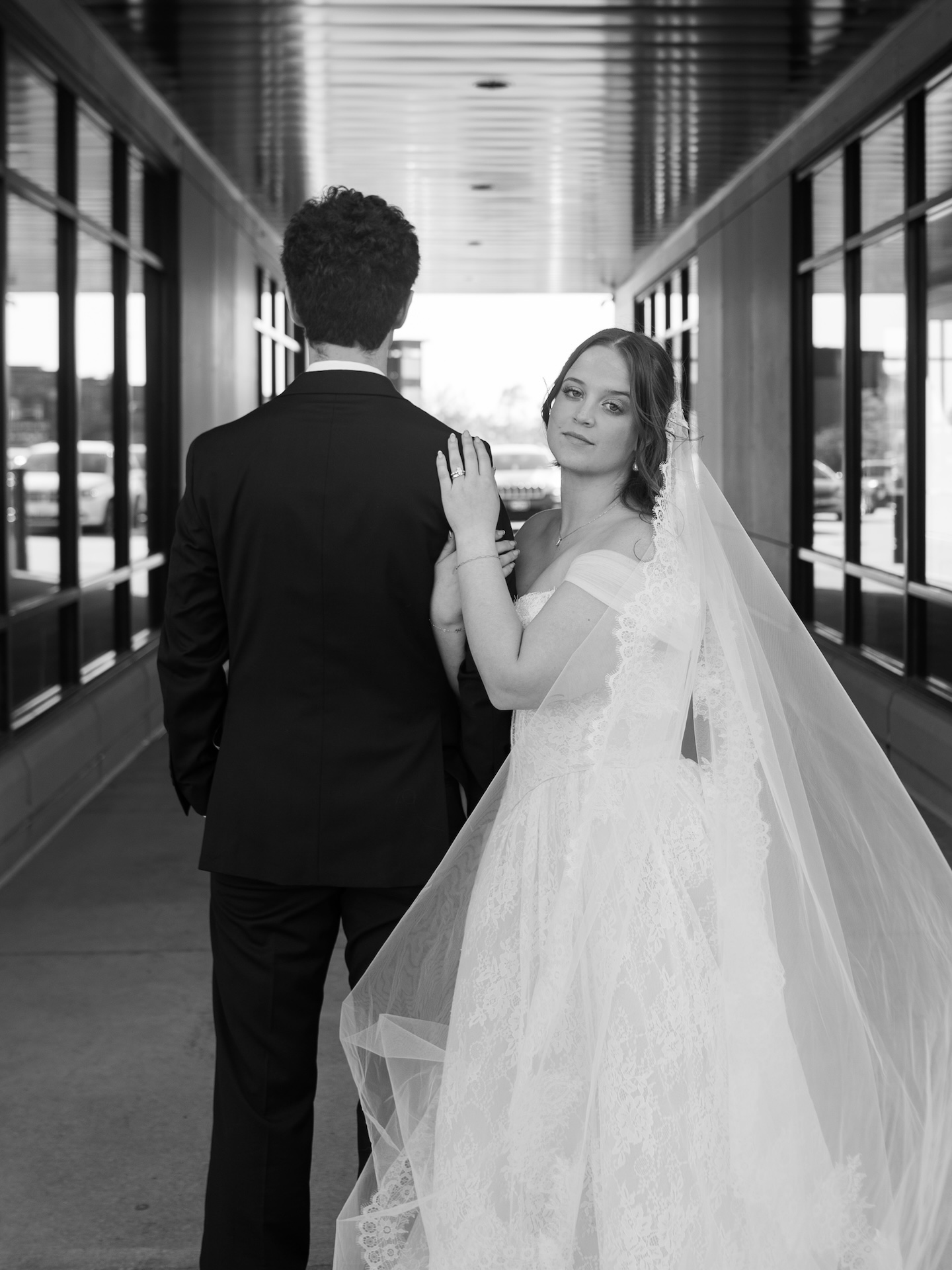 A summer wedding bride in a lace gown and cathedral veil glances back over her shoulder as the groom walks ahead through a corridor
