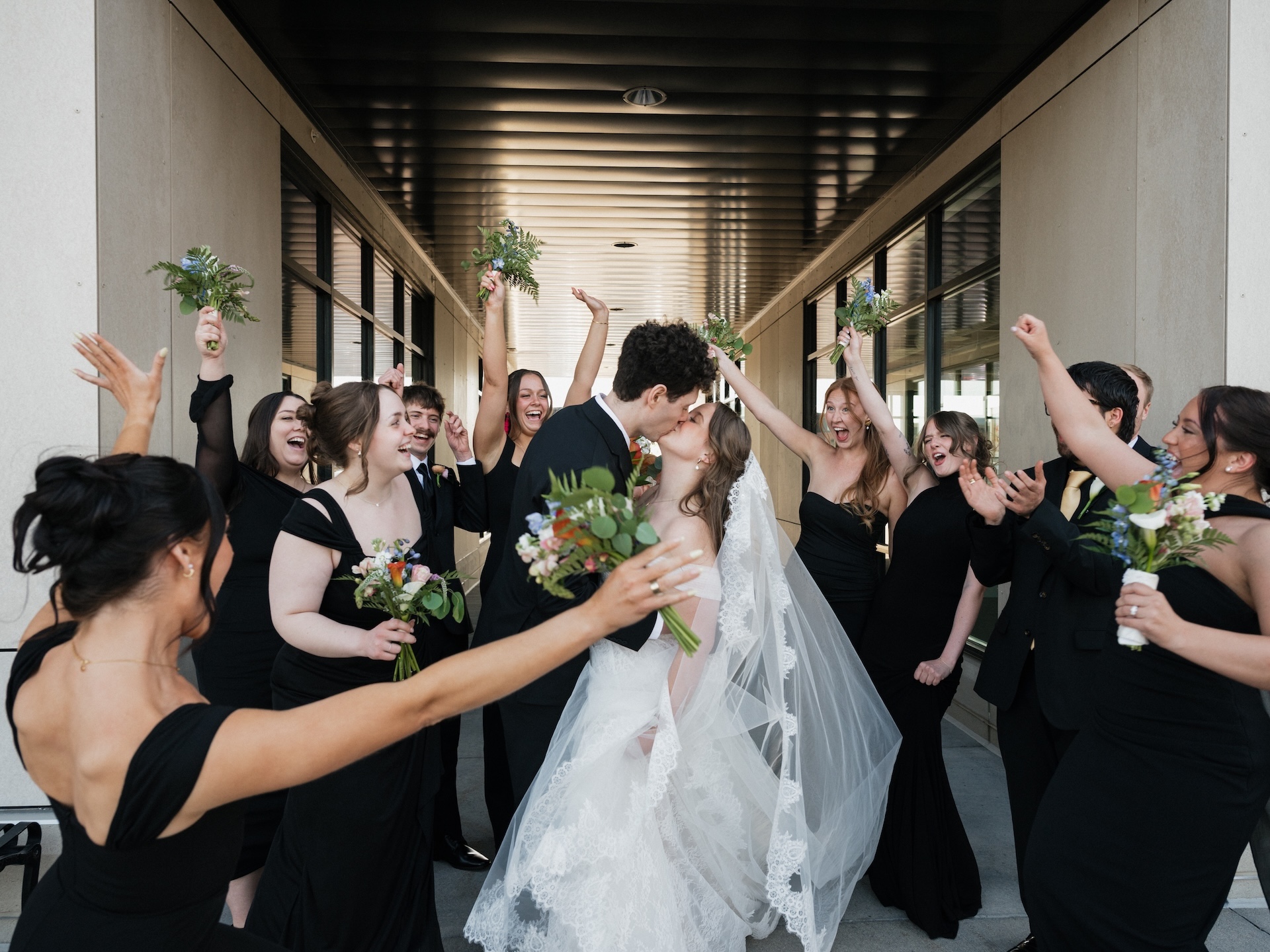 The wedding party surrounds the couple in a corridor, cheering and raising their bouquets as the bride and groom share a kiss
