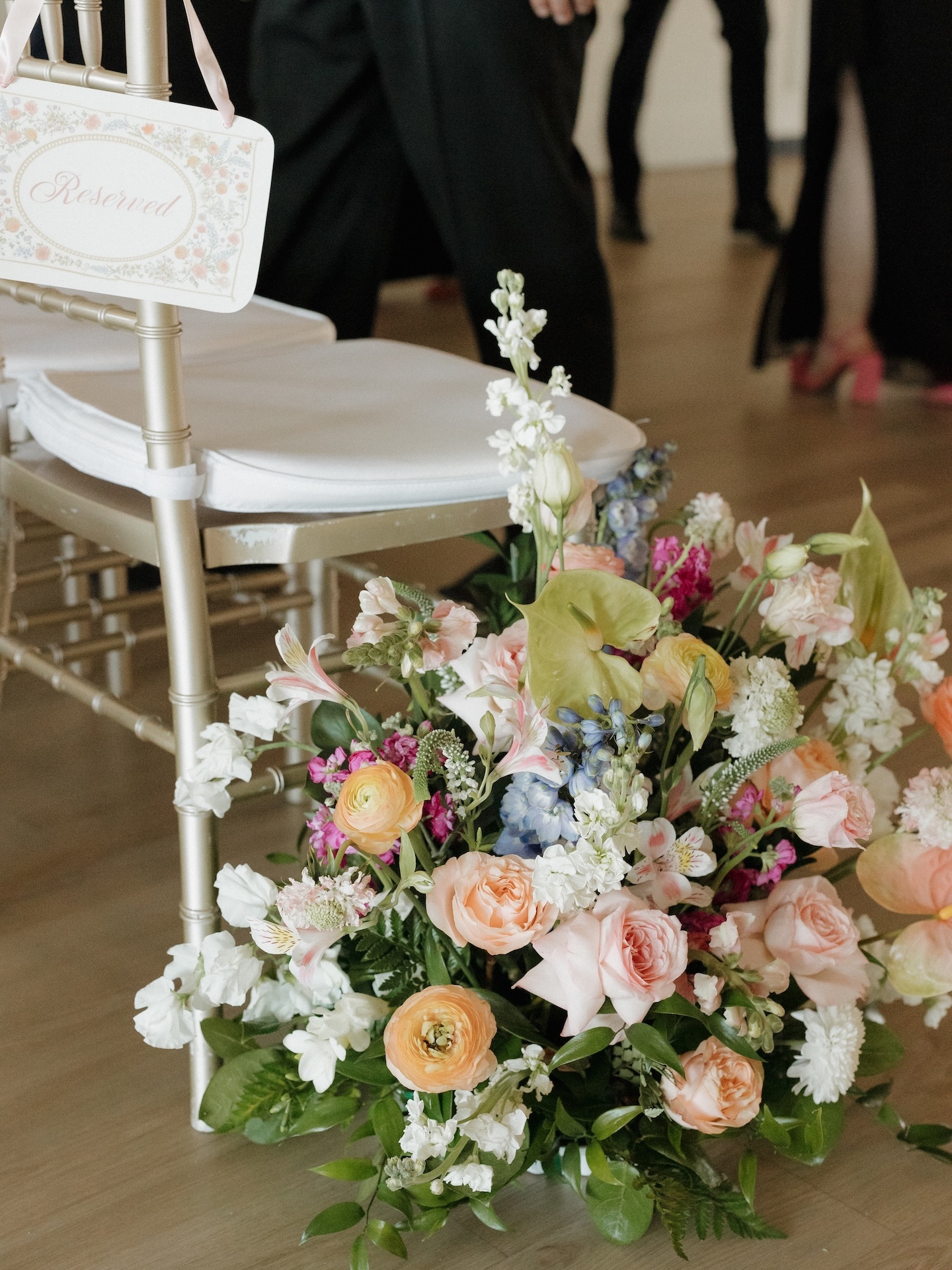 A colorful bridal floor arrangement of peach roses, ranunculus, blue delphinium, and magenta blooms beside a gold chiavari chair with a "Reserved" sign
