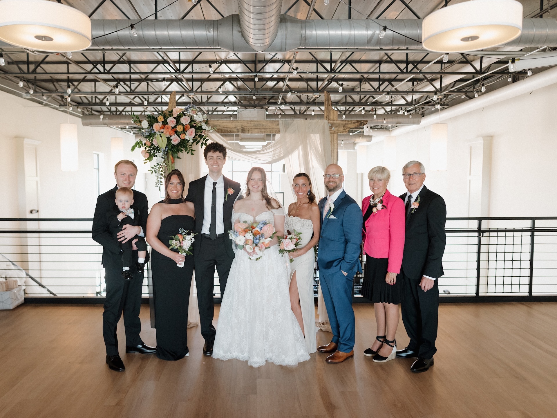 A photo of the bride, groom, and their parents and family surrounded by them in front of their wedding arch.