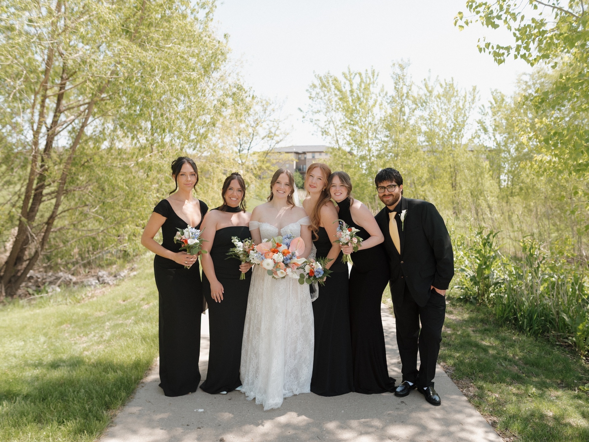 The bride poses with her bridesmaids and a groomsman dressed in black along a tree-lined path on a bright sunny day
