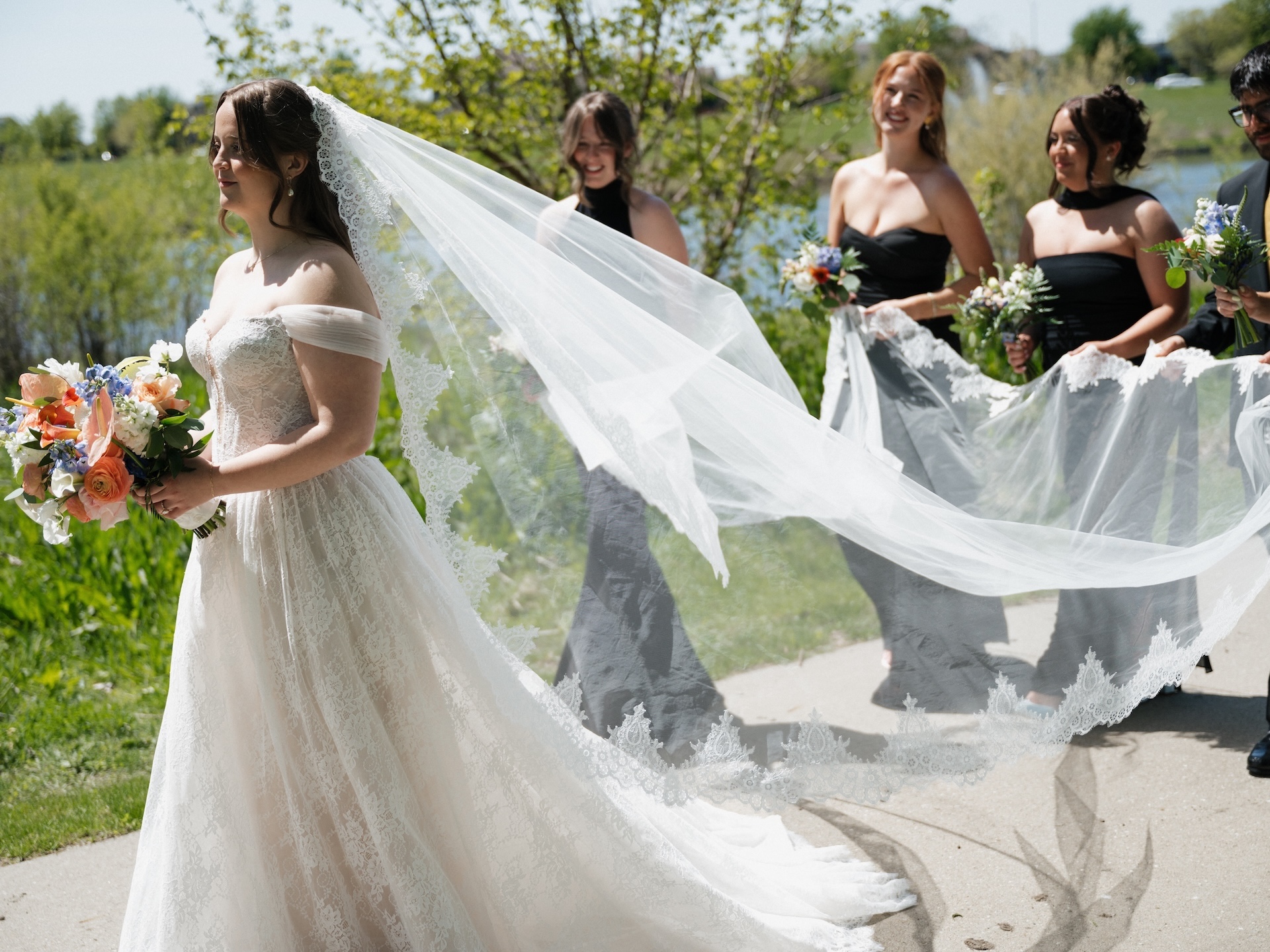 A summer wedding photo of the bride walking ahead as bridesmaids laugh and carry her billowing lace cathedral veil behind her along a waterside path
