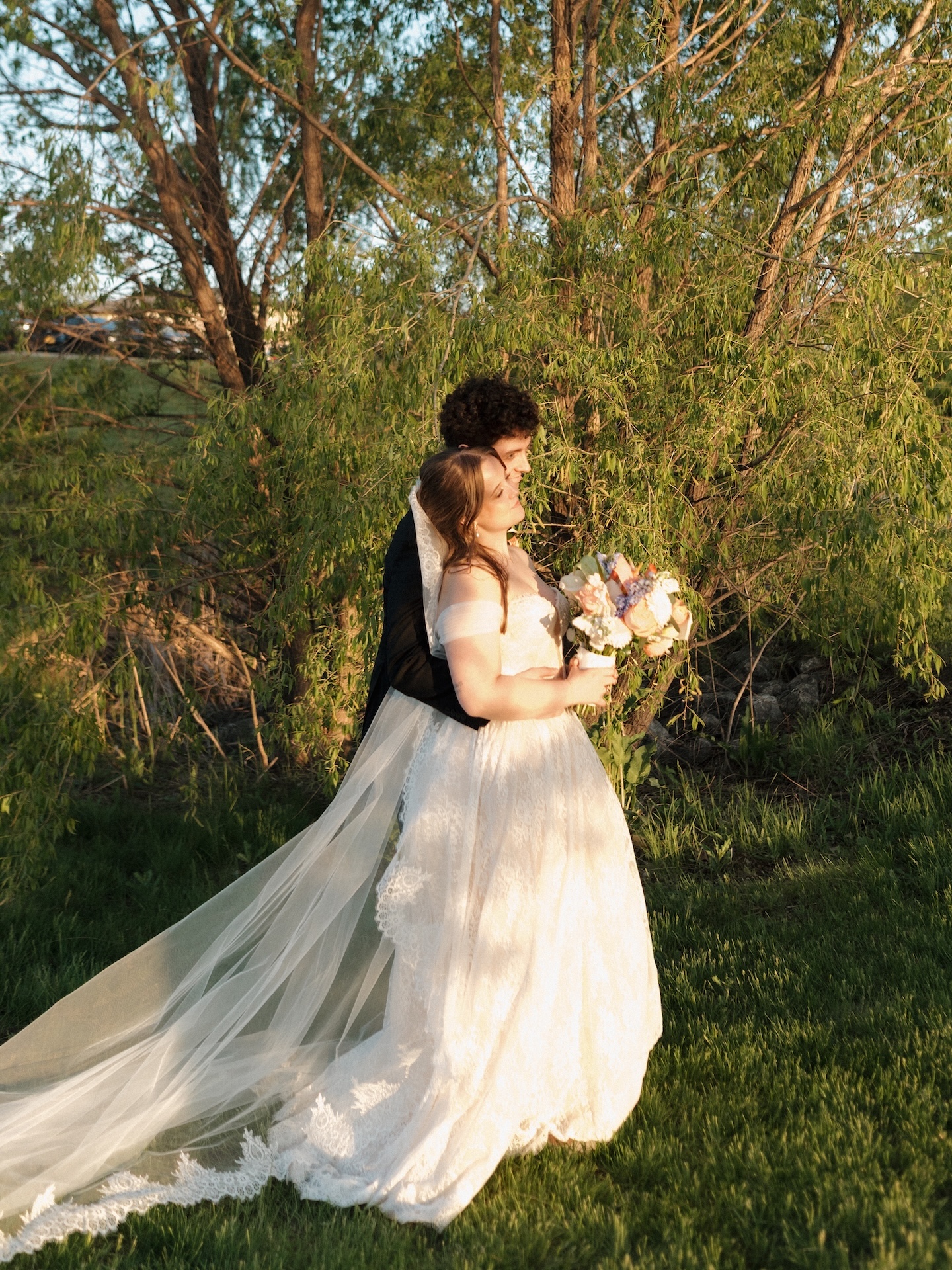 The groom nuzzles the bride from behind as they stand together in front of willow trees during their summer wedding golden hour portraits
