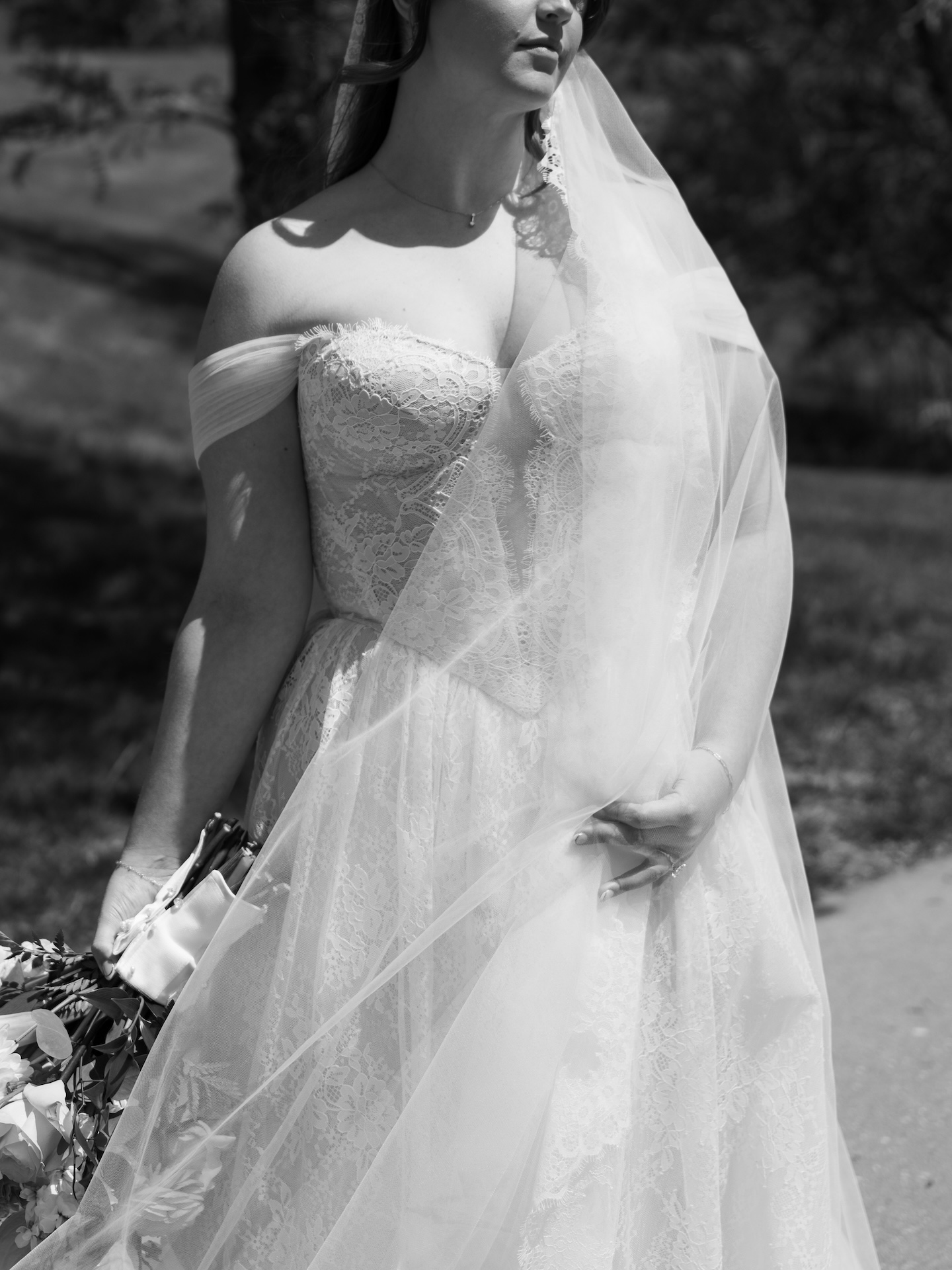 A black and white close-up of the bride's off-the-shoulder lace gown and cathedral veil blowing gently as she holds her bouquet outdoors
