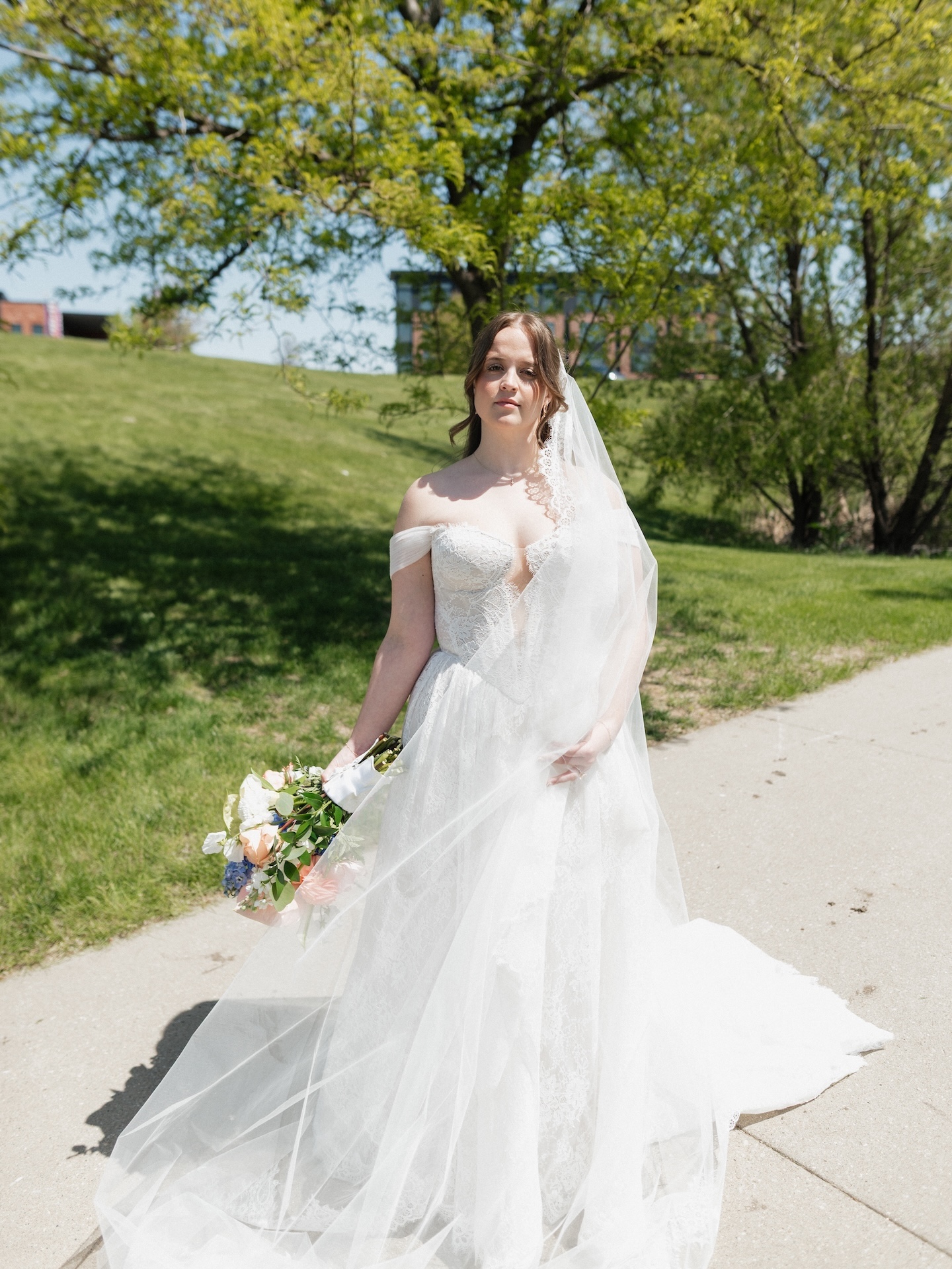 A summer wedding bridal portrait of the bride in her lace off-the-shoulder gown and flowing veil, holding her colorful peach and blue bouquet outdoors
