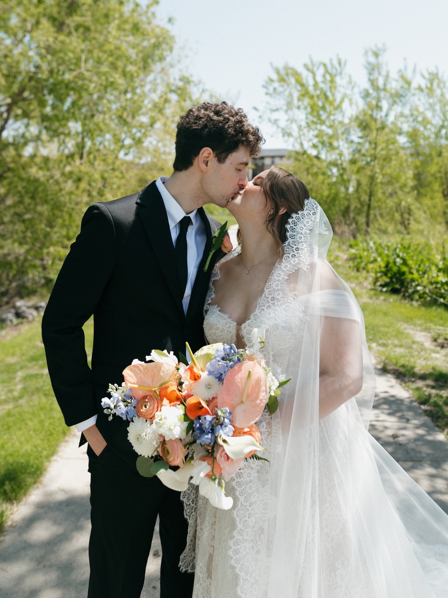 The bride and groom share a kiss outdoors on a sunny spring day, the lace veil catching the breeze while she holds her colorful bouquet
