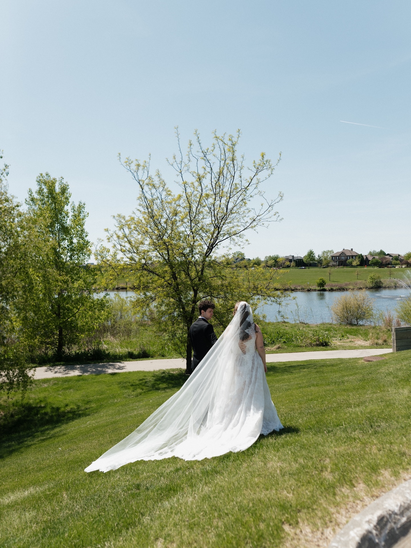 The bride and groom stand together overlooking a peaceful lake on their summer wedding day, the cathedral veil trailing across the green lawn
