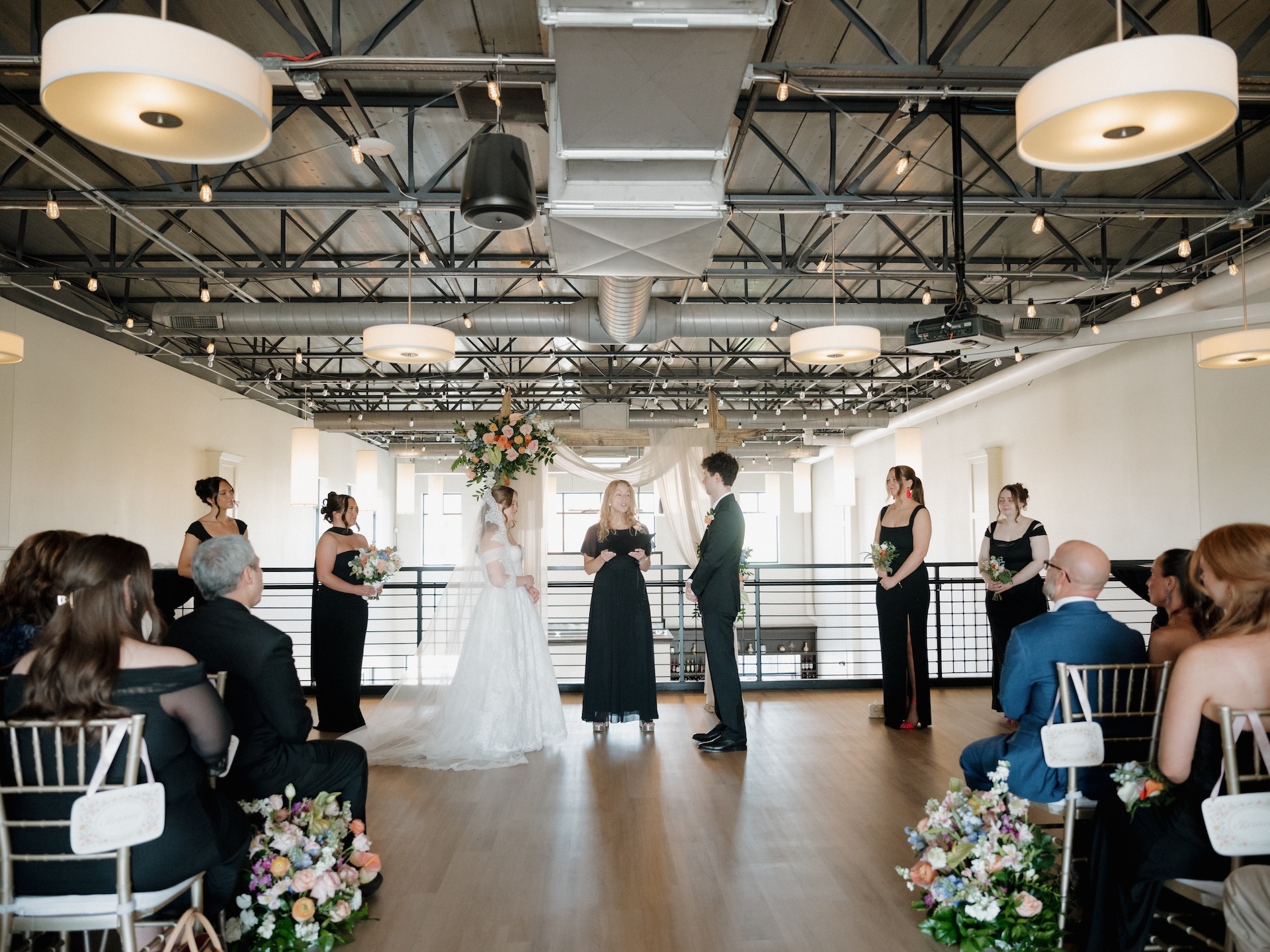 A summer wedding ceremony at The District in Des Moines, Iowa, with the couple exchanging vows beneath a floral arch before seated guests
