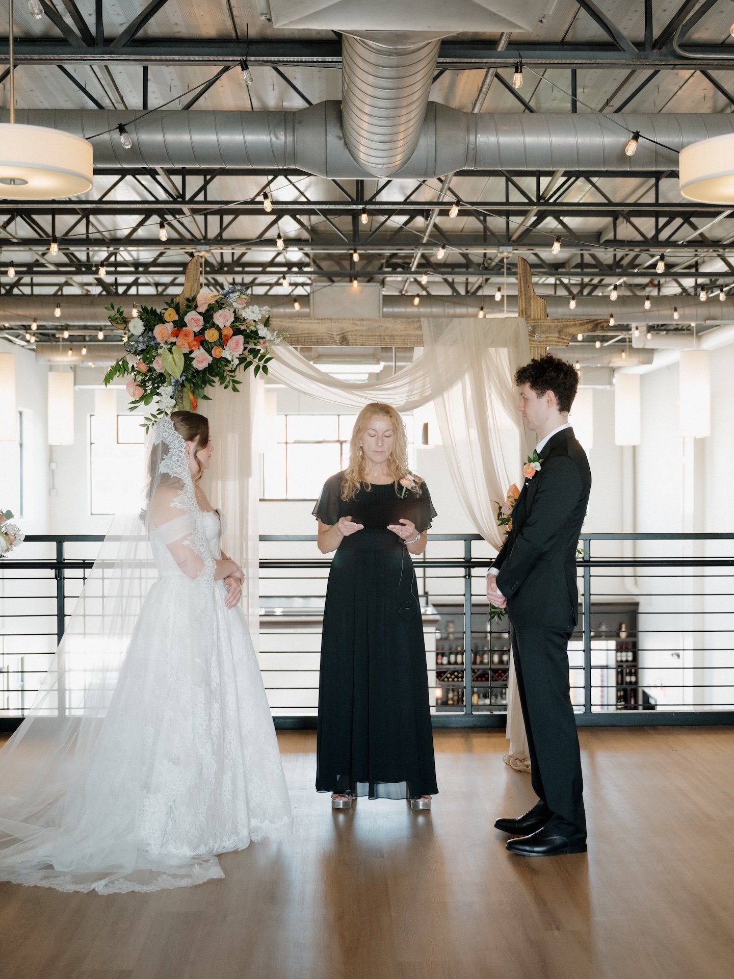 The bride and groom face each other at the altar beneath a lush floral arrangement at The District in Des Moines, Iowa, as the officiant reads
