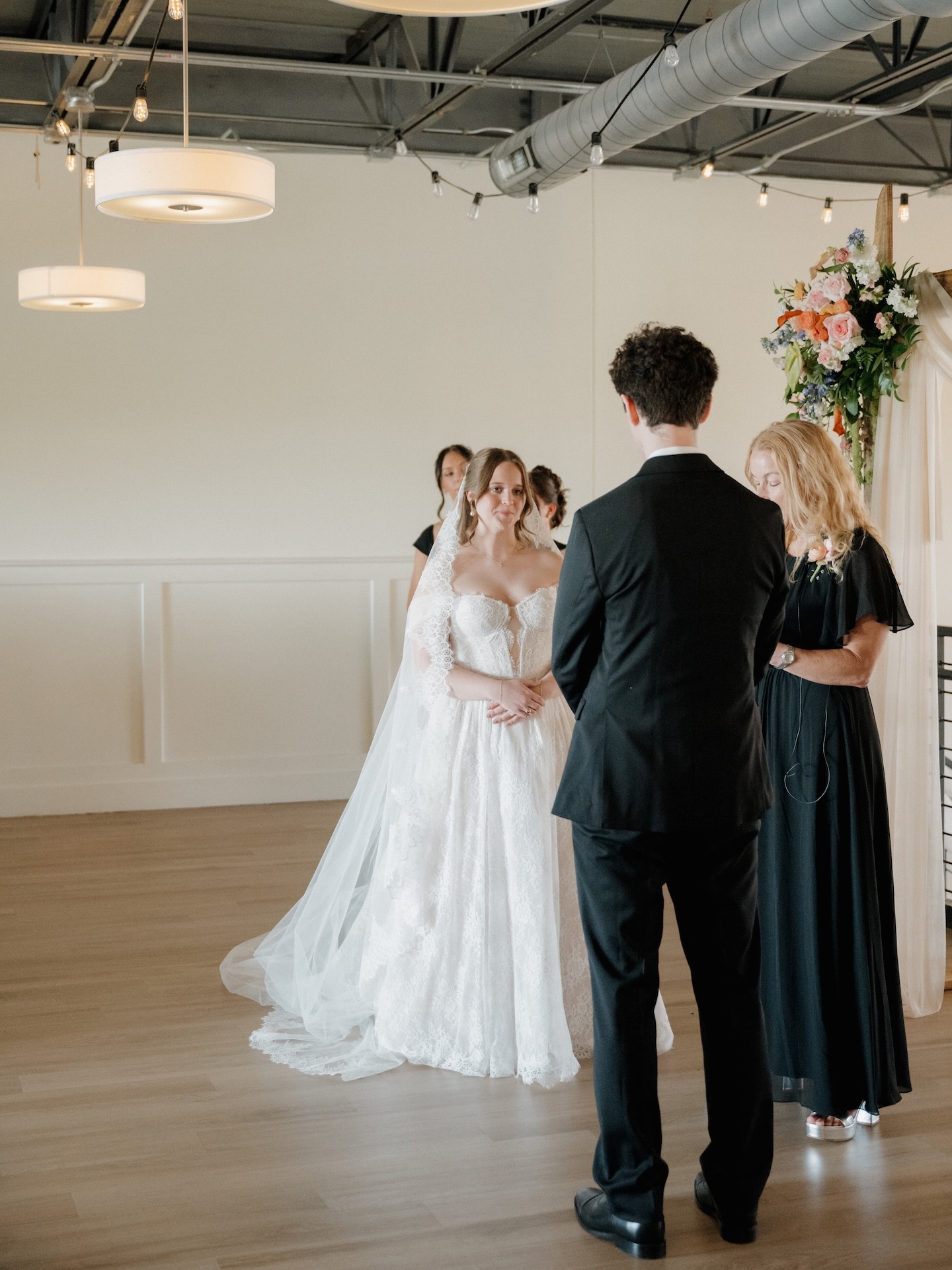 A summer wedding ceremony moment as the bride smiles softly while holding hands with the groom, the officiant reading from a book beside them

