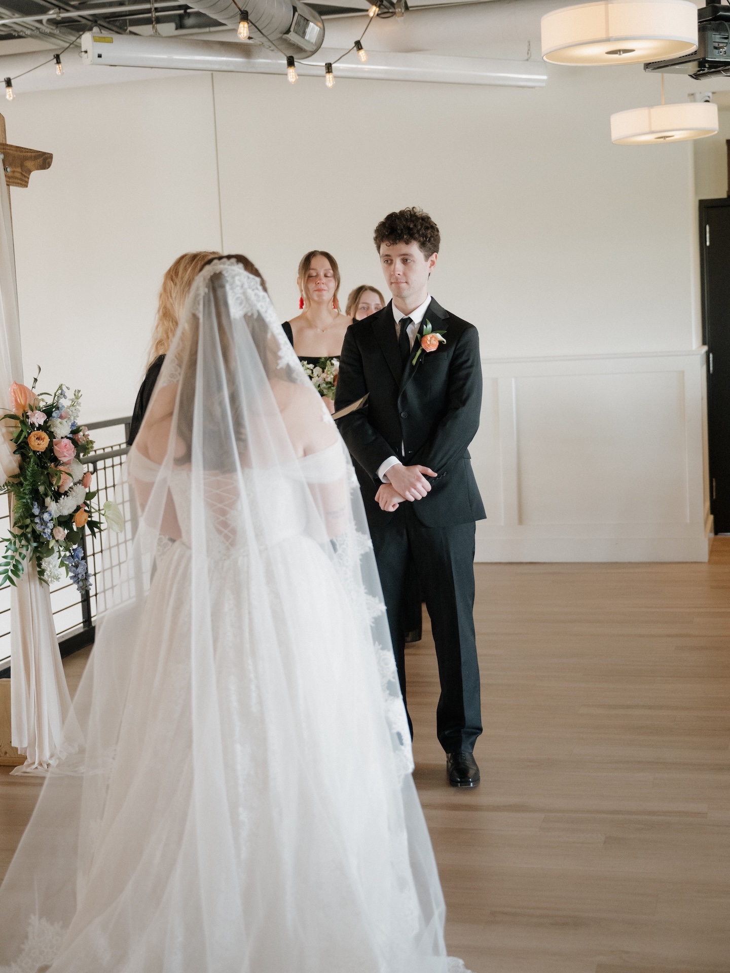 The groom gazes at the bride as she walks toward the altar at The District, a floral arch and draped fabric arch visible in the background
