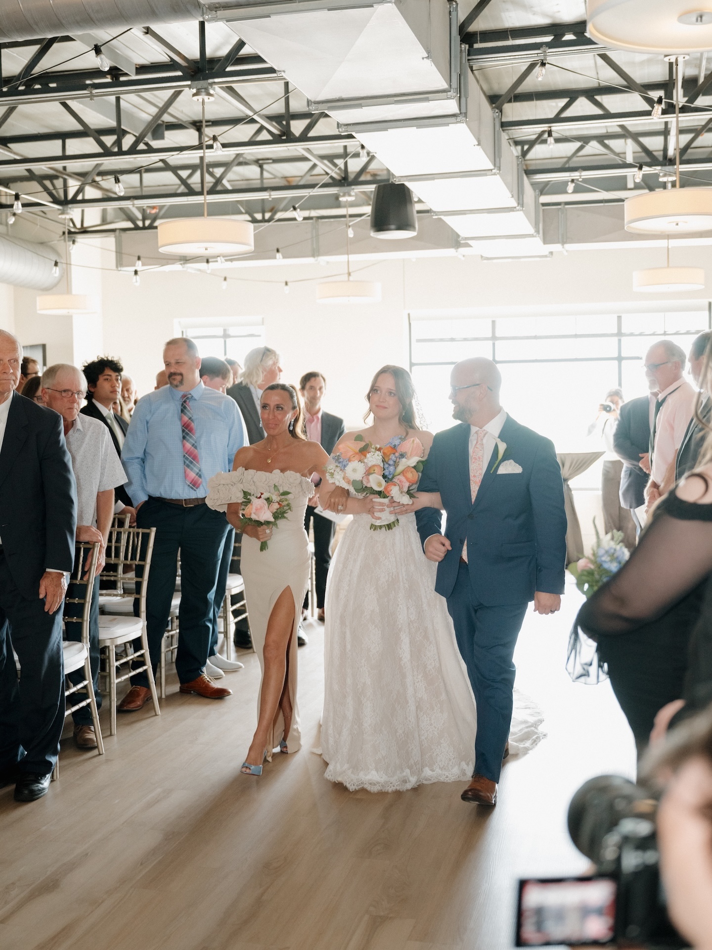 The bride walks down the aisle escorted by both parents during the summer wedding ceremony at The District in Des Moines, Iowa, guests standing on either side
