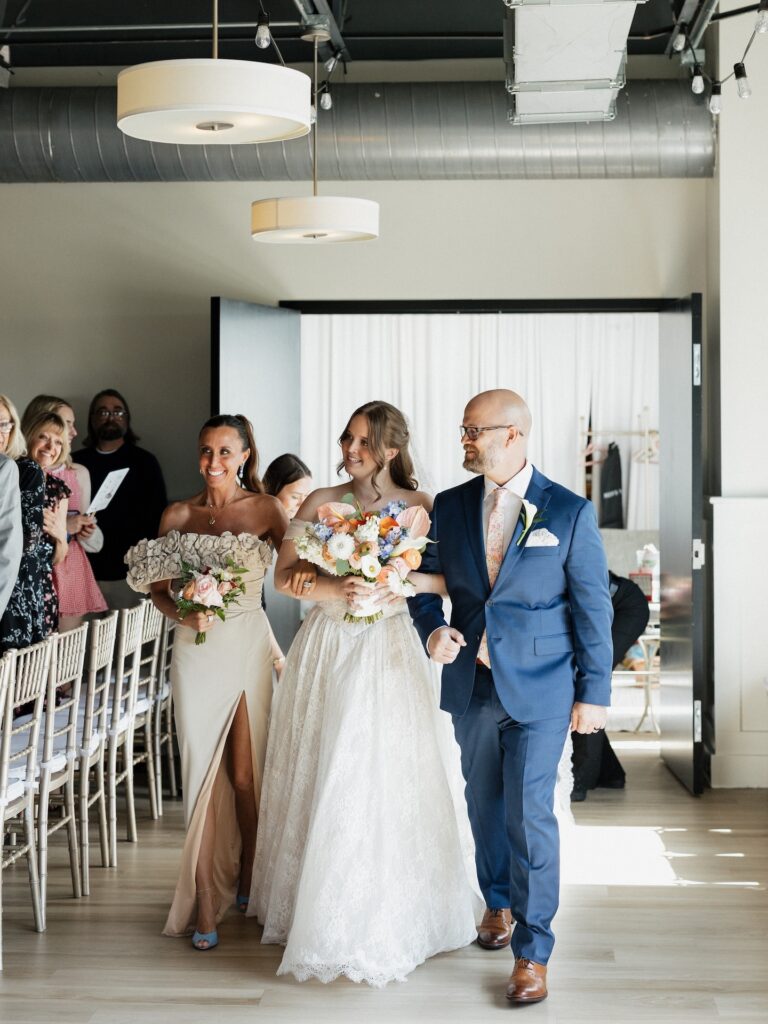 The bride walks down the aisle escorted by both parents, beaming as guests look on during the summer wedding ceremony at The District in Des Moines, Iowa
