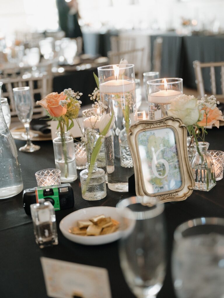 A summer wedding reception tablescape featuring floating candles, bud vases with peach roses, a vintage framed table number, and a disposable camera for guests
