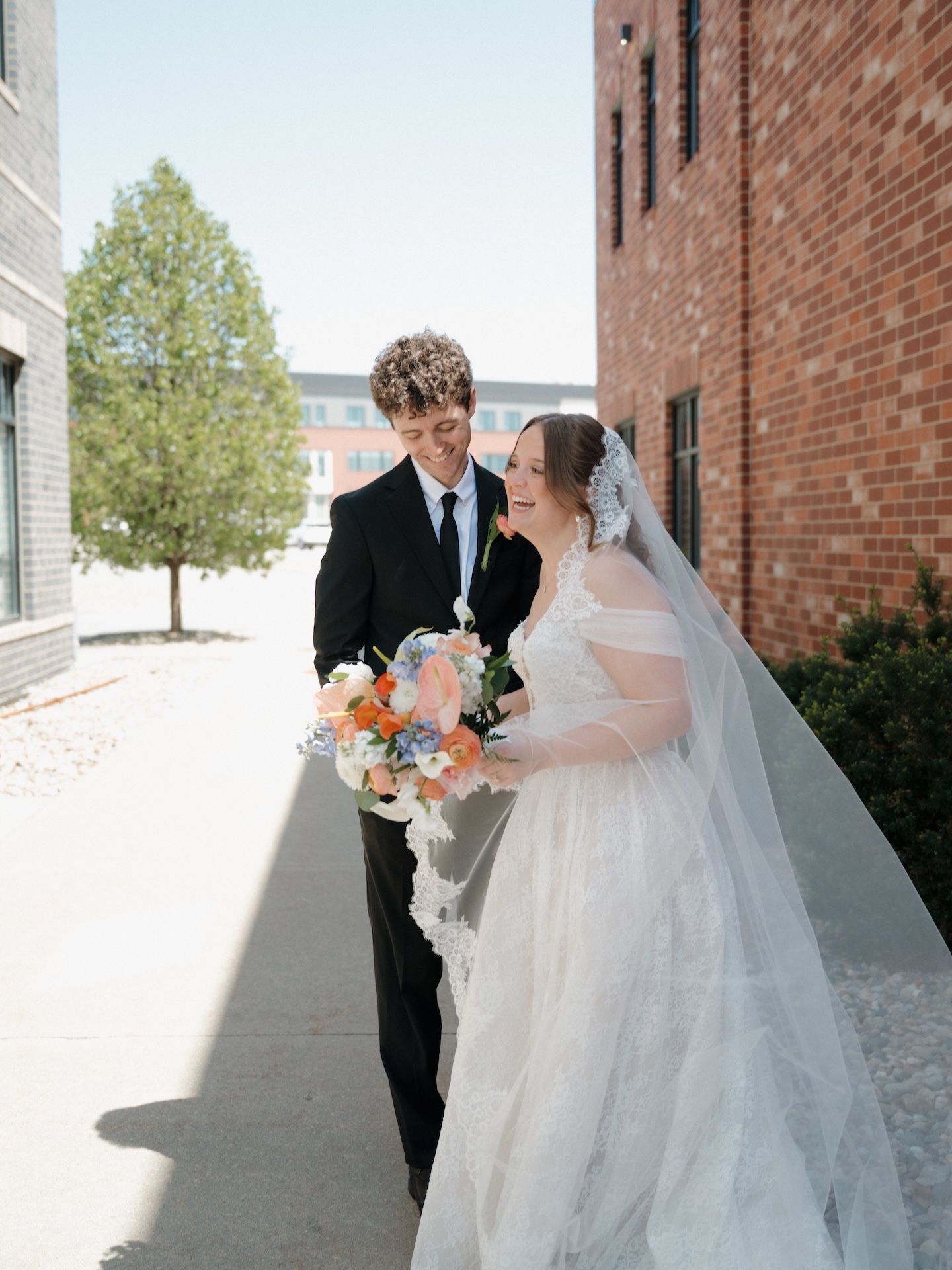 The bride and groom laugh together as they walk along a sunny brick-lined sidewalk, her lace veil catching the light and peach bouquet in hand
