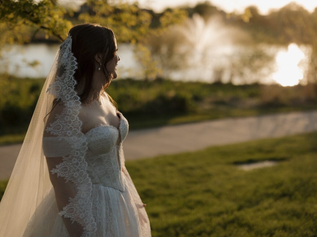 A glowing golden hour bridal portrait of the bride in her lace gown and mantilla veil gazing toward a sunlit fountain and lake at dusk
