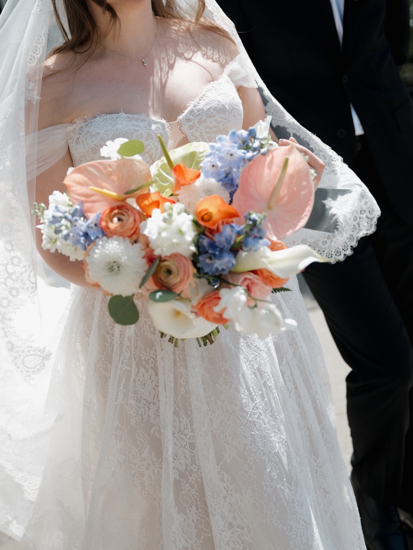 A close-up of the bride holding her vibrant summer wedding bouquet of peach anthuriums, orange ranunculus, blue hydrangea, white calla lilies, and eucalyptus
