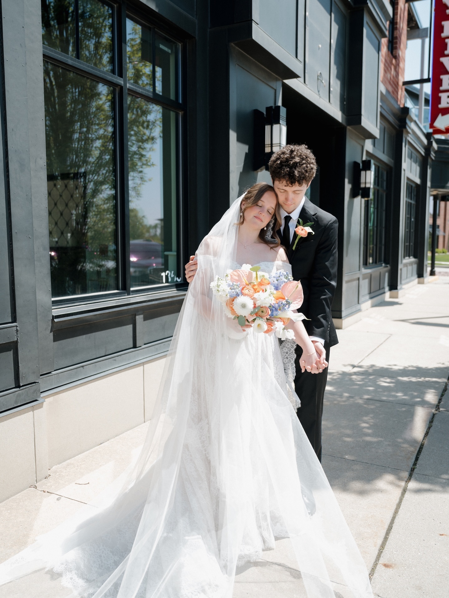 A summer wedding couple embraces on a city sidewalk, the bride resting her head on the groom's shoulder while holding a vibrant peach and blue bouquet
